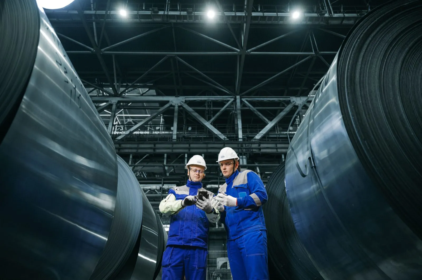 Deux techniciens en casque entre des bobines d'acier dans une halle industrielle